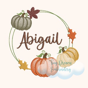 Könnte beinhalten: Ein Stickmuster zum Thema Herbst mit einem Kranz aus drei Kürbissen und Blättern sowie dem Namen "Abigail" in brauner Kursivschrift.