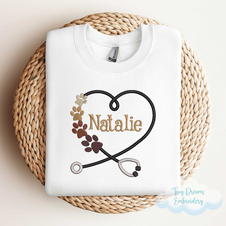 May include: White sweatshirt with a brown and black paw print heart design and the name "Natalie" embroidered in gold. A stethoscope is crossed over the heart. The text "Tiny Dreams Embroidery" is in the bottom right corner.