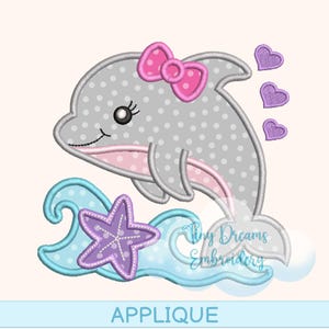 May include: An applique design featuring a smiling gray dolphin with a pink bow, polka dots, and a pink belly. The dolphin is above blue waves, a purple starfish, and three purple hearts. The word "APPLIQUE" is at the bottom.