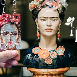 May include: A pair of beaded earrings featuring a portrait of Frida Kahlo. The earrings are handmade and have a red, white, and blue color scheme. The earrings are hanging from a silver hook.