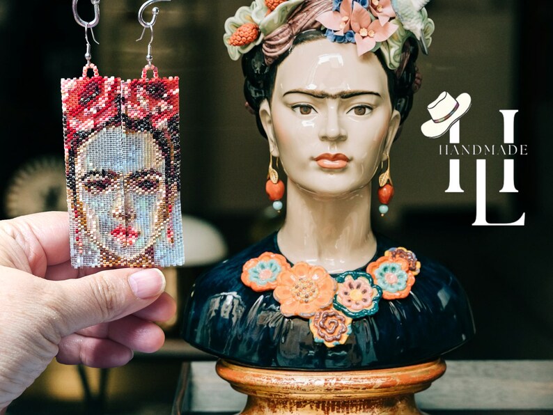 May include: A pair of beaded earrings featuring a portrait of Frida Kahlo. The earrings are handmade and have a red, white, and blue color scheme. The earrings are hanging from a silver hook.