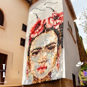 May include: A pair of handmade beaded earrings featuring a portrait of Frida Kahlo with red flowers in her hair. The earrings are hanging on a white wall.