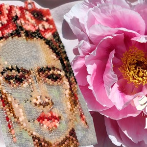 May include: A beaded portrait of Frida Kahlo with a pink peony flower in the background. The portrait is made with colorful beads and the flower is in full bloom.
