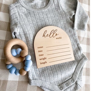 May include: A wooden baby announcement plaque with the text "hello world" and blank lines for name, date, time, weight, and length. The plaque is on a grey baby onesie with a wooden rattle.