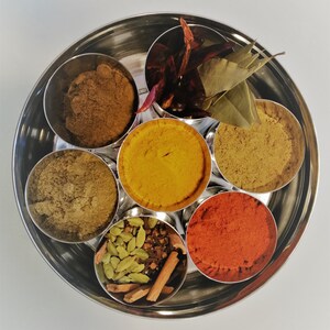 Op de afbeelding: Een ronde metalen container met zeven compartimenten, elk gevuld met een ander specerij. De specerijen omvatten kurkuma, chilipoeder, komijn, koriander, kardemom, kaneelstokjes en laurierbladeren.