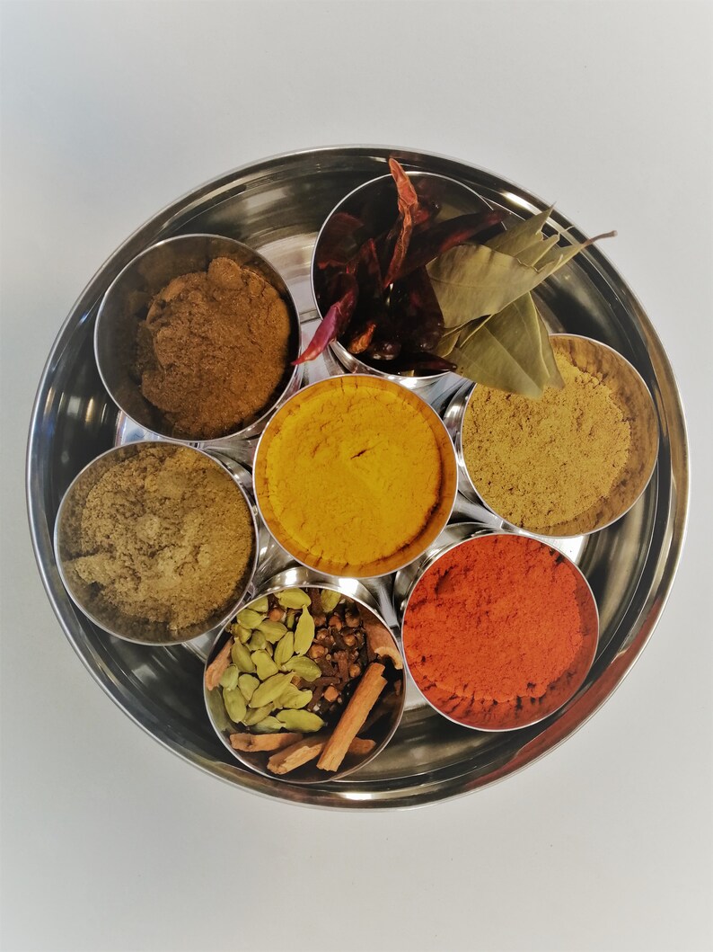 Op de afbeelding: Een ronde metalen container met zeven compartimenten, elk gevuld met een ander specerij. De specerijen omvatten kurkuma, chilipoeder, komijn, koriander, kardemom, kaneelstokjes en laurierbladeren.