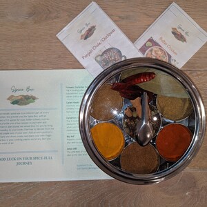 Op de afbeelding: Een roestvrijstalen specerijenbox met een doorzichtig deksel, met zes compartimenten gevuld met verschillende specerijen. De doos staat op een houten oppervlak met twee receptenkaarten voor Punjabi Chole/Kikkererwten en Botersauskip.