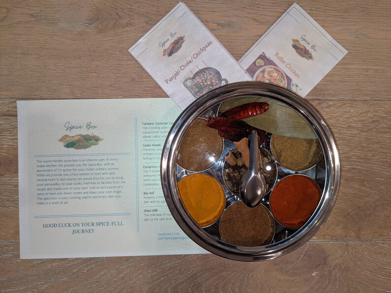 Op de afbeelding: Een roestvrijstalen specerijenbox met een doorzichtig deksel, met zes compartimenten gevuld met verschillende specerijen. De doos staat op een houten oppervlak met twee receptenkaarten voor Punjabi Chole/Kikkererwten en Botersauskip.