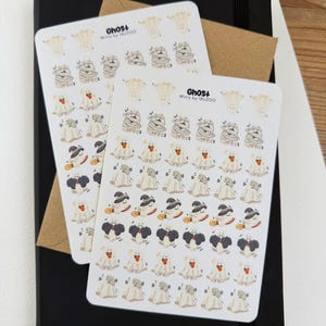 May include: Two sticker sheets featuring Halloween-themed ghost illustrations in various costumes, including mummies and witches. The stickers are on a black notebook with a brown envelope. The text "Ghost+ Minis by IBUZOO" is visible on the stickers.