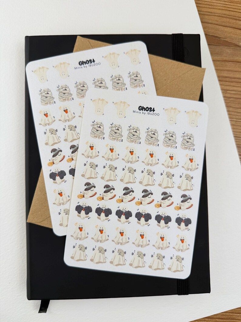 May include: Two sticker sheets featuring Halloween-themed ghost illustrations in various costumes, including mummies and witches. The stickers are on a black notebook with a brown envelope. The text "Ghost+ Minis by IBUZOO" is visible on the stickers.