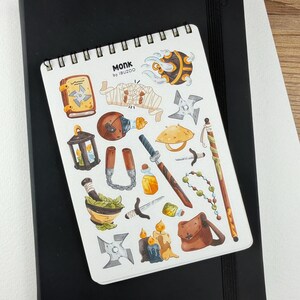 Op de afbeelding: Een spiraalgebonden notitieblok met een verzameling ge&iuml;llustreerde stickers met een monnikenthema. De stickers beelden verschillende items uit, waaronder een rol, wapens en een tas. Het notitieblok ligt op een zwart notitieboek.