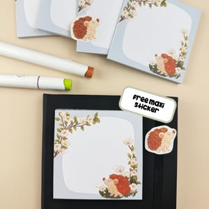 Può includere: Blocchi per appunti azzurri con disegni floreali agli angoli e un'illustrazione di riccio dei cartoni animati. Sono presenti anche un taccuino nero, pennarelli bianchi e un'etichetta "Free maxi sticker". Sono visibili anche adesivi a forma di riccio.