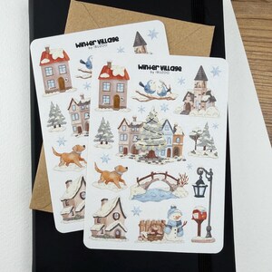 May include: Two white sticker sheets titled Winter Village by IBUZOO featuring watercolor illustrations of snow-covered houses, a decorated Christmas tree, a golden dog, a stone bridge, a snowman, a street lamp, and blue snowflakes on a black notebook.