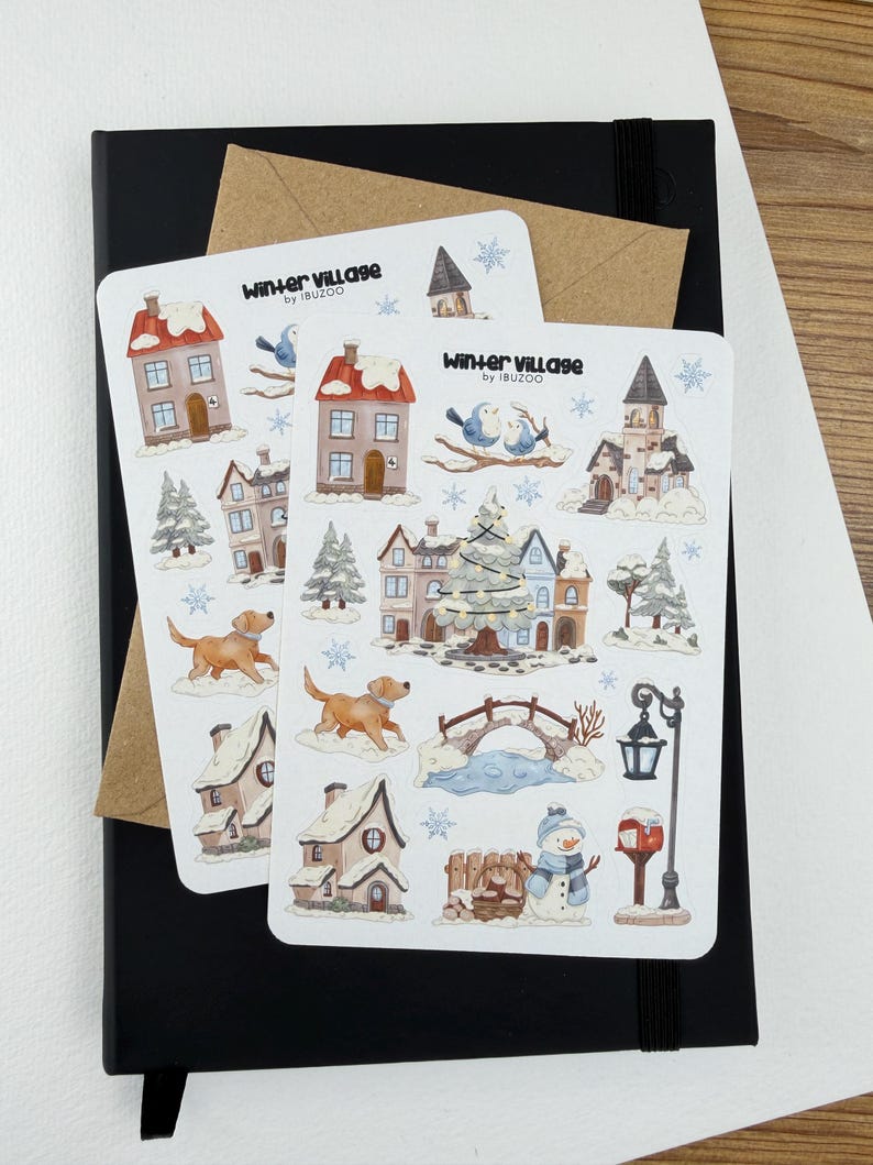 May include: Two white sticker sheets titled Winter Village by IBUZOO featuring watercolor illustrations of snow-covered houses, a decorated Christmas tree, a golden dog, a stone bridge, a snowman, a street lamp, and blue snowflakes on a black notebook.