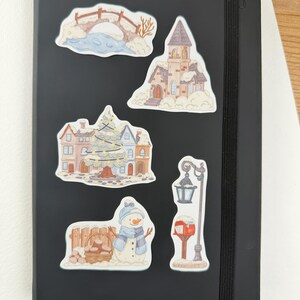 May include: A black notebook featuring five winter-themed stickers: a snow-covered bridge, a tall church, a town square with a decorated Christmas tree, a snowman standing next to a wooden fence with a basket of logs, and a street lamp with a mailbox.
