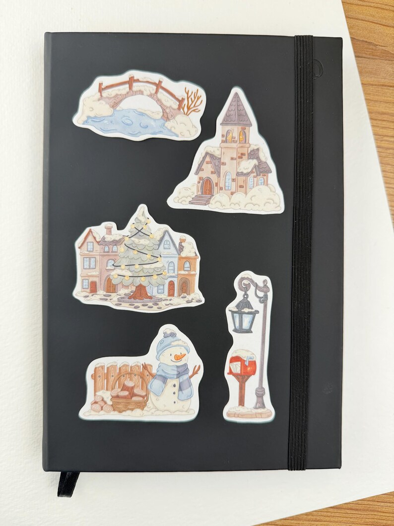 May include: A black notebook featuring five winter-themed stickers: a snow-covered bridge, a tall church, a town square with a decorated Christmas tree, a snowman standing next to a wooden fence with a basket of logs, and a street lamp with a mailbox.