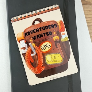 Op de afbeelding: Een spiraalgebonden notitieblok met een cartoonillustratie van een bruine rugzak met de tekst "ADVENTURERS WANTED" en "RPG". Het notitieblok ligt bovenop een zwart notitieboek. Het notitieblok heeft ook een stickervel label met "20 Stickersheets".