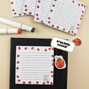 Könnte beinhalten: Weiße Notizblöcke mit schwarzen Linien und roten Marienkäfer-Illustrationen. Die Notizblöcke tragen den Text "LUCKY NOTES". Ein weißer Marker und ein "free maxi sticker" mit Marienkäfer-Design sind ebenfalls vorhanden.