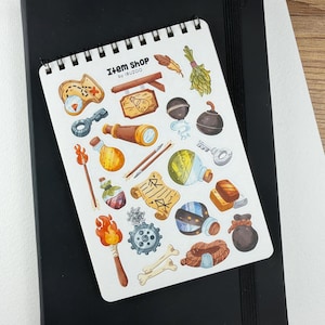 Op de afbeelding: Een spiraalgebonden notitieblok met diverse stickers met fantasiethema's, waaronder een kaart, drankflessen, een telescoop en een fakkel. Het notitieblok is gemarkeerd met "Item Shop by IBUZOO" en ligt op een zwart notitieboek.
