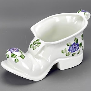 May include: A white ceramic shoe-shaped planter with a handle and floral design. The planter has a heel and toe, with purple and green floral accents. The interior is hollow, and the exterior is glossy.