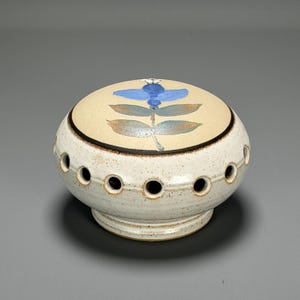 May include: A ceramic container with a beige lid featuring a blue floral design. The container has a speckled white body with evenly spaced holes around the middle. A black band encircles the lid's edge.