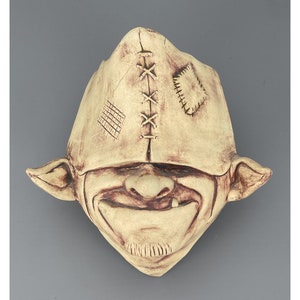 May include: A ceramic goblin mask with a stitched-on hat and a toothy grin. The mask is painted in shades of brown and beige.