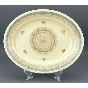 May include: A large, oval, cream-coloured serving platter with a delicate, lace-like design in shades of green and brown. The centre of the platter features a circular design with a radial pattern.