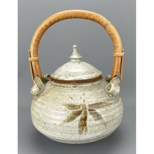 May include: A ceramic teapot with a light beige speckled finish and a dragonfly design. The teapot has a bamboo handle and a matching lid with a finial. The teapot is a decorative kitchenware item.