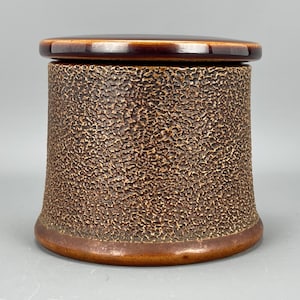 May include: A brown ceramic jar with a textured surface and a lid. The jar has a rounded shape and a slightly flared lip.