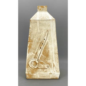 May include: A tall, off-white ceramic vase with a rectangular base and a tapered top. The vase features a raised relief design of a pair of scissors. The vase has a textured surface and a rustic, handmade appearance.