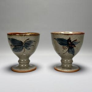 May include: Two ceramic goblets with a speckled grey finish. Each goblet features a dark blue butterfly or floral design. The rims and bases have a reddish-brown trim. The goblets are on a pedestal base.