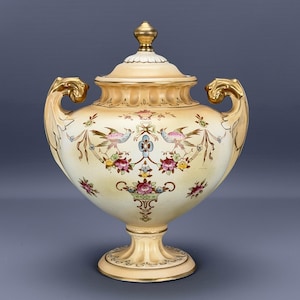 May include: A cream-coloured, lidded urn with gold accents and handles. The urn is decorated with floral and bird motifs in pink, blue, and yellow. The base and lid are also cream-coloured with gold trim.