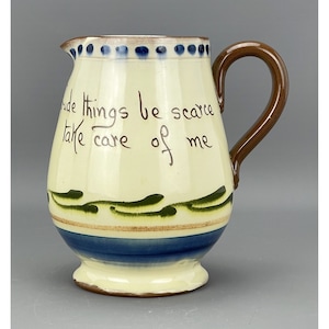 May include: A cream-coloured ceramic jug with a brown handle. It has a blue and green design with the text "Rude things be scarce take care of me".