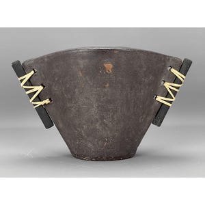 May include: A dark brown bowl with a distinctive form. It has black wooden handles on each side, fastened with cream-coloured twine. The bowl's exterior has a textured appearance, suggesting a natural material. The bowl is approximately 20 cm in diameter.