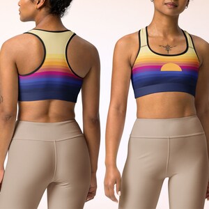 May include: A sports bra with a sunset graphic print. The bra has a racerback style and is made of a stretchy, moisture-wicking fabric. The fabric is a light yellow color with a multi-colored sunset graphic print. The bra has a black band around the top and bottom.