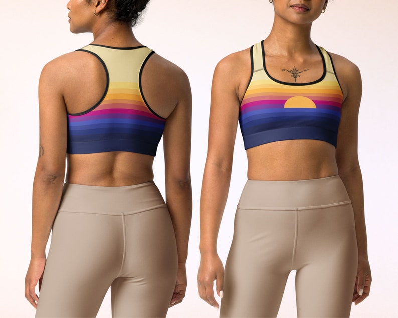 May include: A sports bra with a sunset graphic print. The bra has a racerback style and is made of a stretchy, moisture-wicking fabric. The fabric is a light yellow color with a multi-colored sunset graphic print. The bra has a black band around the top and bottom.