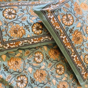 Könnte beinhalten: Bettwäsche-Set mit floralem Muster. Das Design zeigt große orangefarbene und gelbe Blumen mit grünen Blättern auf hellblauem Hintergrund. Die Kissenbezüge und das Laken haben einen dekorativen Rand. Die Website-Adresse "https://ploutus.biz" ist sichtbar.