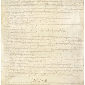 Puede incluir: Un primer plano de un documento hist&oacute;rico, la Constituci&oacute;n de los Estados Unidos, escrita en letra cursiva en papel envejecido. El texto incluye la frase "Art&iacute;culo II" y el comienzo de la secci&oacute;n 1. El documento es una pieza significativa de la historia estadounidense.