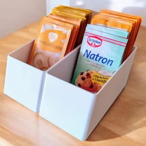 Puede incluir: Dos contenedores rectangulares blancos con tapas, uno lleno de pequeños paquetes naranjas etiquetados "Vanillinzucker" y el otro lleno de pequeños paquetes azules y naranjas etiquetados "Dr. Oetker Natron".