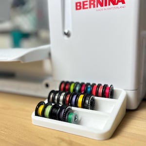 May include: White plastic thread organizer with multiple spools of colorful thread. The organizer has three rows and is sitting on a wooden surface. The text "BERNINA MADE IN SWITZERLAND" is visible on the white sewing machine in the background.