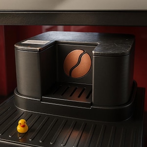 Peut inclure: Machine à expresso noire avec un motif de grain de café en cuivre. La machine a une surface texturée et un petit canard en caoutchouc jaune est sur le plateau. La machine est sur une surface noire.