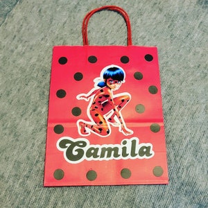 May include: Red and black polka dot gift bag with a cartoon image of a superheroine with black hair and a red and black costume. The bag has the name "Camila" printed on the front.