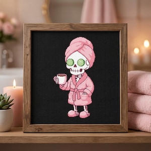 May include: A square wooden frame containing a cross-stitch pattern on a black background. The design features a white skeleton wearing a pink bathrobe, a matching pink towel wrapped around its head, and green cucumber slices over its eyes.