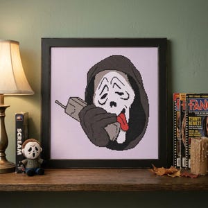 May include: A framed cross-stitch artwork featuring a pixelated Ghostface character wearing a dark hooded robe, holding a gray telephone handset to their ear, and sticking out a bright red tongue, set against a light purple grid background.