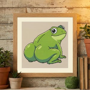 May include: A framed cross-stitch artwork featuring a bright green frog with large eyes and a rounded, prominent posterior. The frog is depicted in a seated position against a light beige background, enclosed within a light-toned wooden frame.