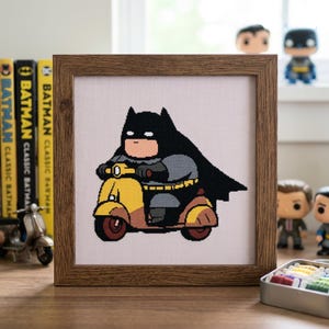May include: A framed cross-stitch embroidery featuring a stylized, rounded Batman character riding a yellow and brown scooter. The character wears a black cape and mask, set against a white grid fabric background within a dark wood-toned frame.