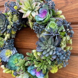 May include: A green and blue succulent wreath with purple flowers. The wreath is hanging on a wooden door.