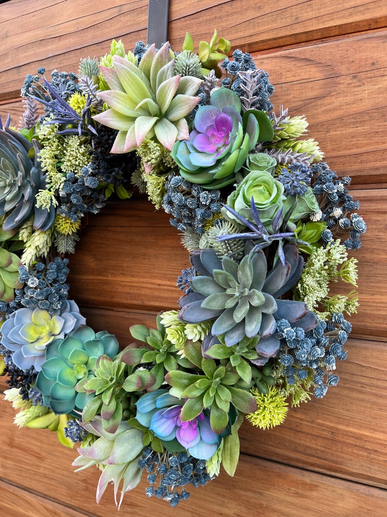 May include: A green and blue succulent wreath with purple flowers. The wreath is hanging on a wooden door.