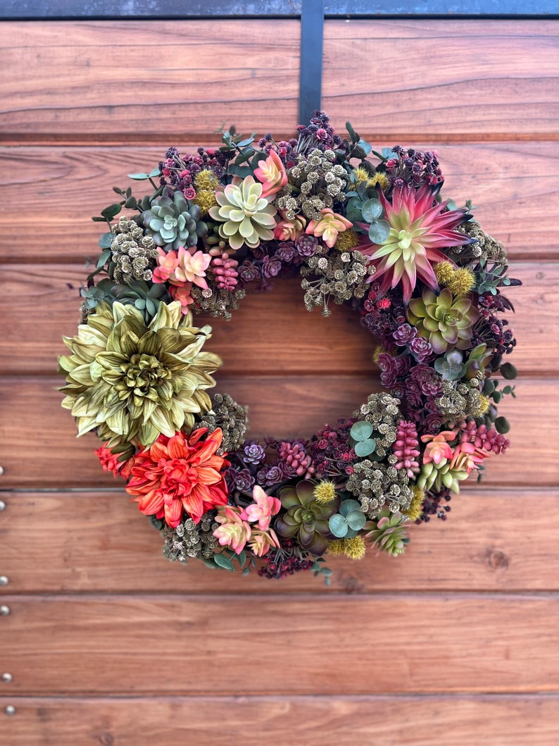 May include: A green, red, and pink succulent wreath with a variety of textures and colors. The wreath is hanging on a wooden door.