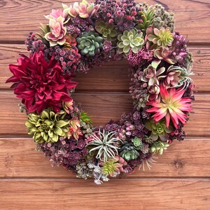 May include: A green and burgundy succulent wreath with a variety of succulents and burgundy flowers. The wreath is hanging on a wooden door.